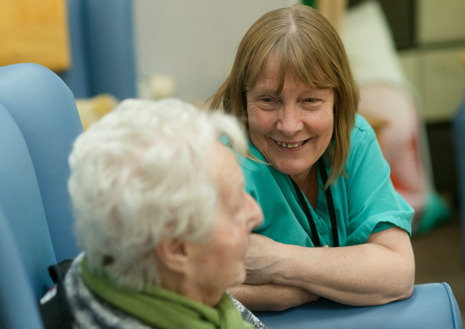 A healthcare worker in a teal shirt with a stethoscope smiles while interacting with an elderly patient with white hair in what appears to be a clinical setting.