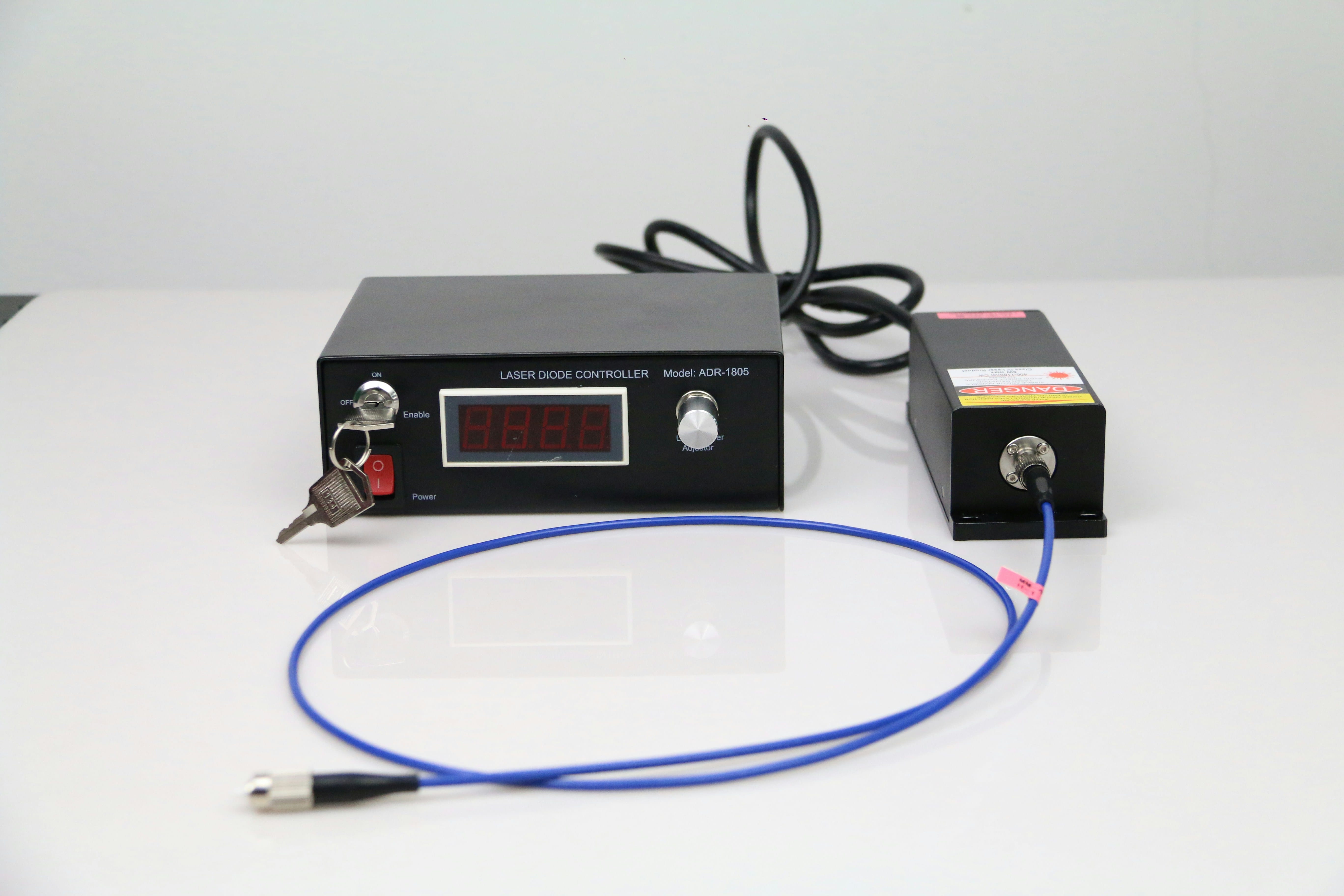 A dark laboratory instrument with a digital display panel and control knobs sits on a white surface next to a smaller gold-colored box, with a blue cable looped in front of them and a black power cord visible.