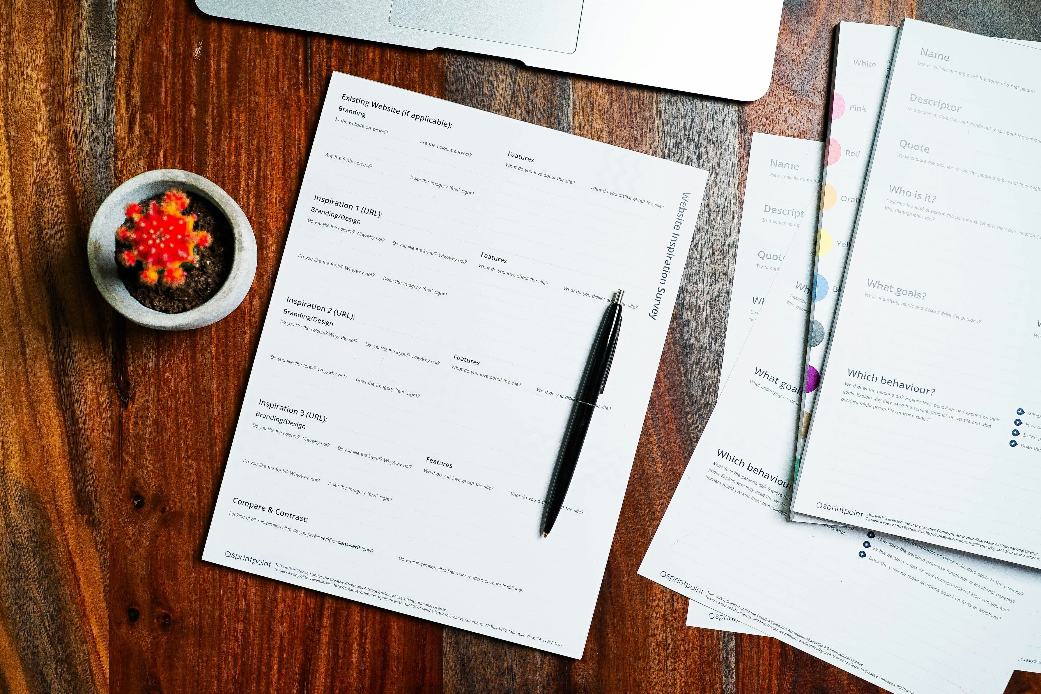 A wooden desk surface displaying multiple printed business documents with lined pages, a black pen, a purple pen, and a small white bowl containing red flowers, photographed from above in landscape orientation.