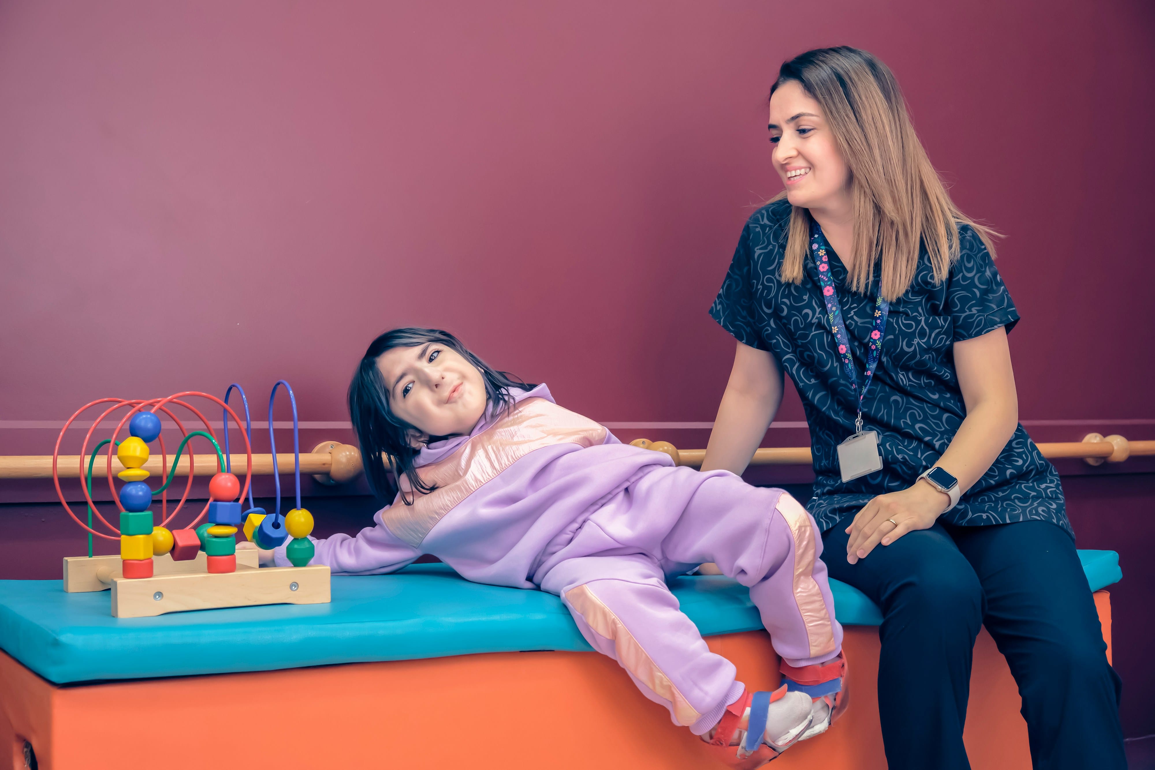 An adult woman and a young child sit together on colorful foam platforms against a pink wall, with the child lying back while the woman looks at them supportively; a toy sensory bead maze is visible beside them, suggesting a playful therapeutic or counseling environment.