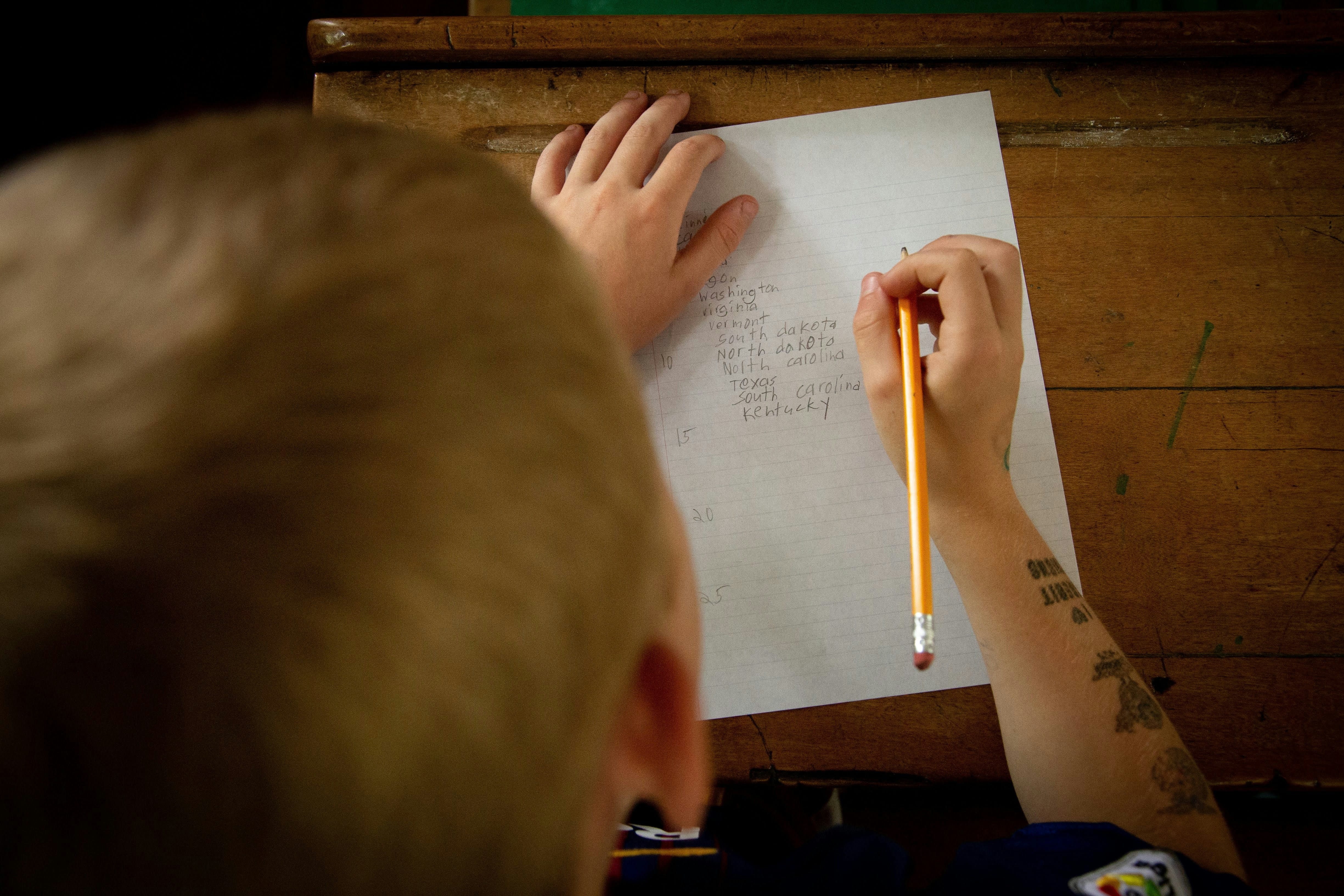 A person with a tattoo on their arm sits at a wooden desk holding a pencil over a white sheet of paper, appearing to write or draw, with another hand visible adjusting the paper.
