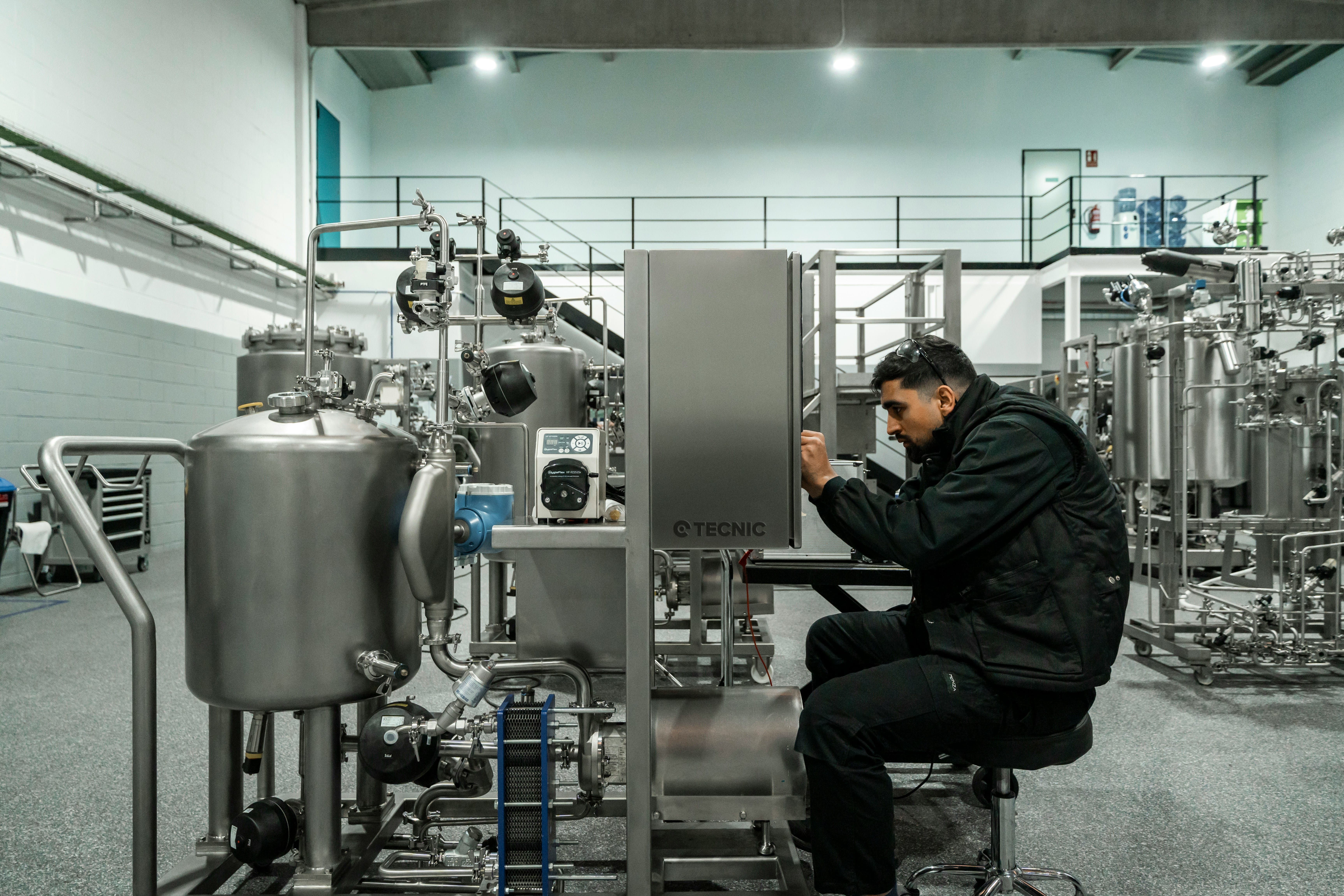A technician in dark clothing crouches while inspecting large stainless steel industrial equipment and piping systems in a modern manufacturing facility with metal railings and overhead structures visible in the background.