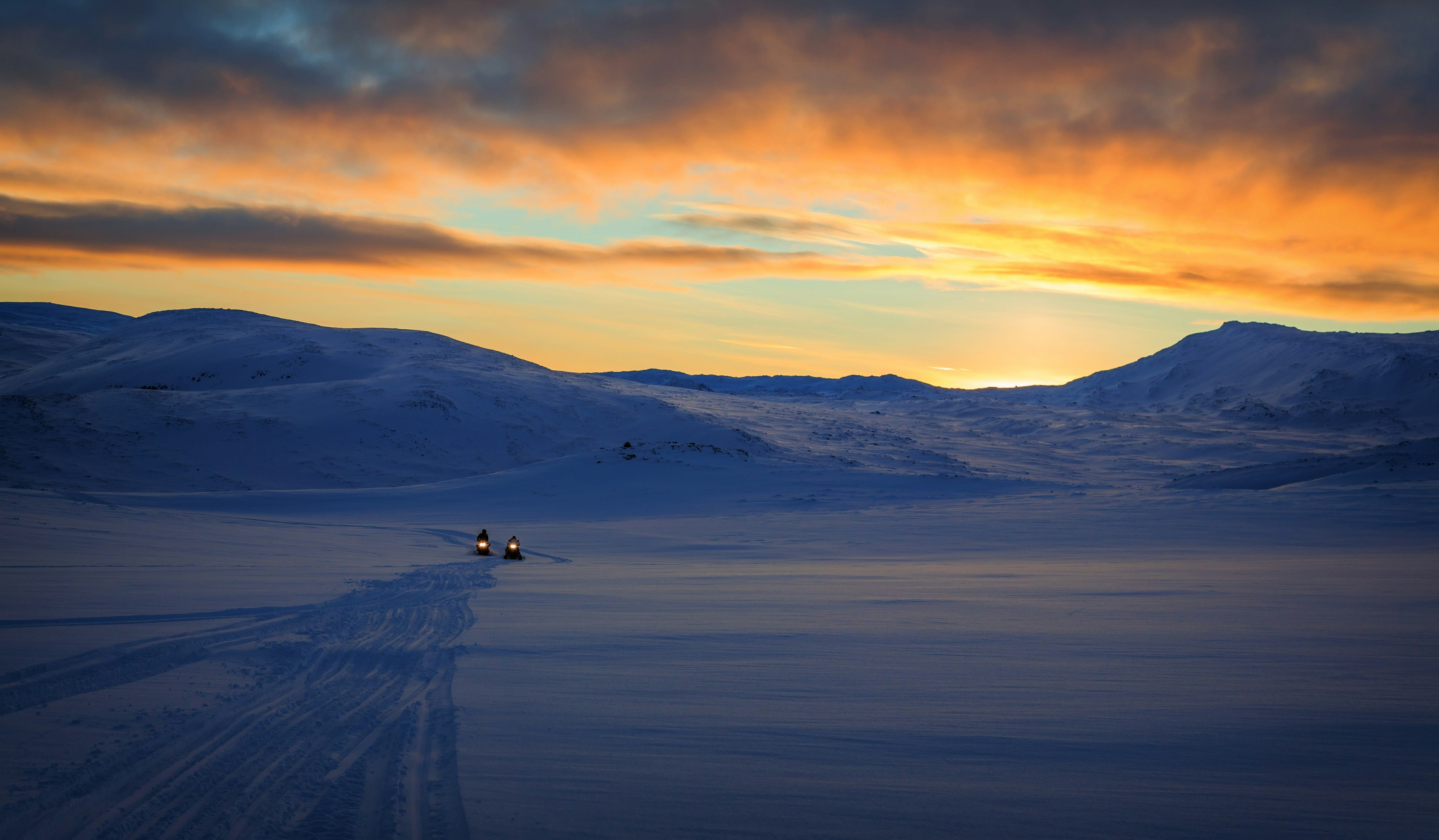 A frozen lake landscape at twilight with golden and orange sky reflected on ice, mountains visible in the distance under a dramatic cloudy horizon, and a small structure or vehicle visible on the frozen surface.